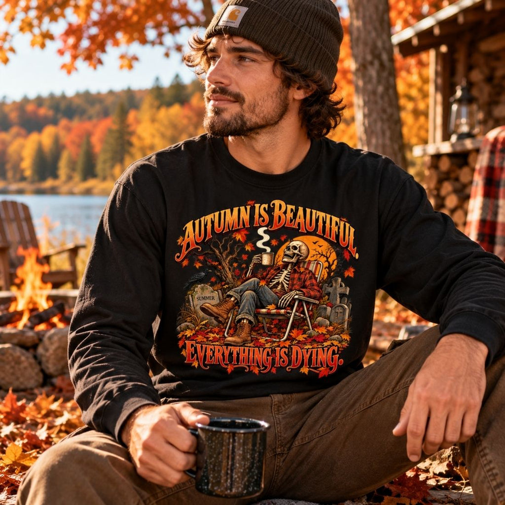 Model wearing a black Autumn Is Beautiful long-sleeve T-shirt and brown pants, seated outdoors with a mug amid fall foliage.