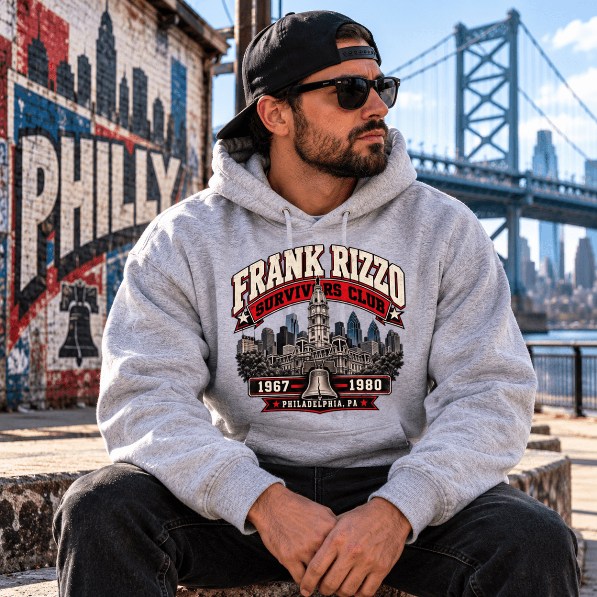 Model wearing a light gray Frank Rizzo Survivors Club hoodie, dark pants, sunglasses and backward cap, seated near a bridge.