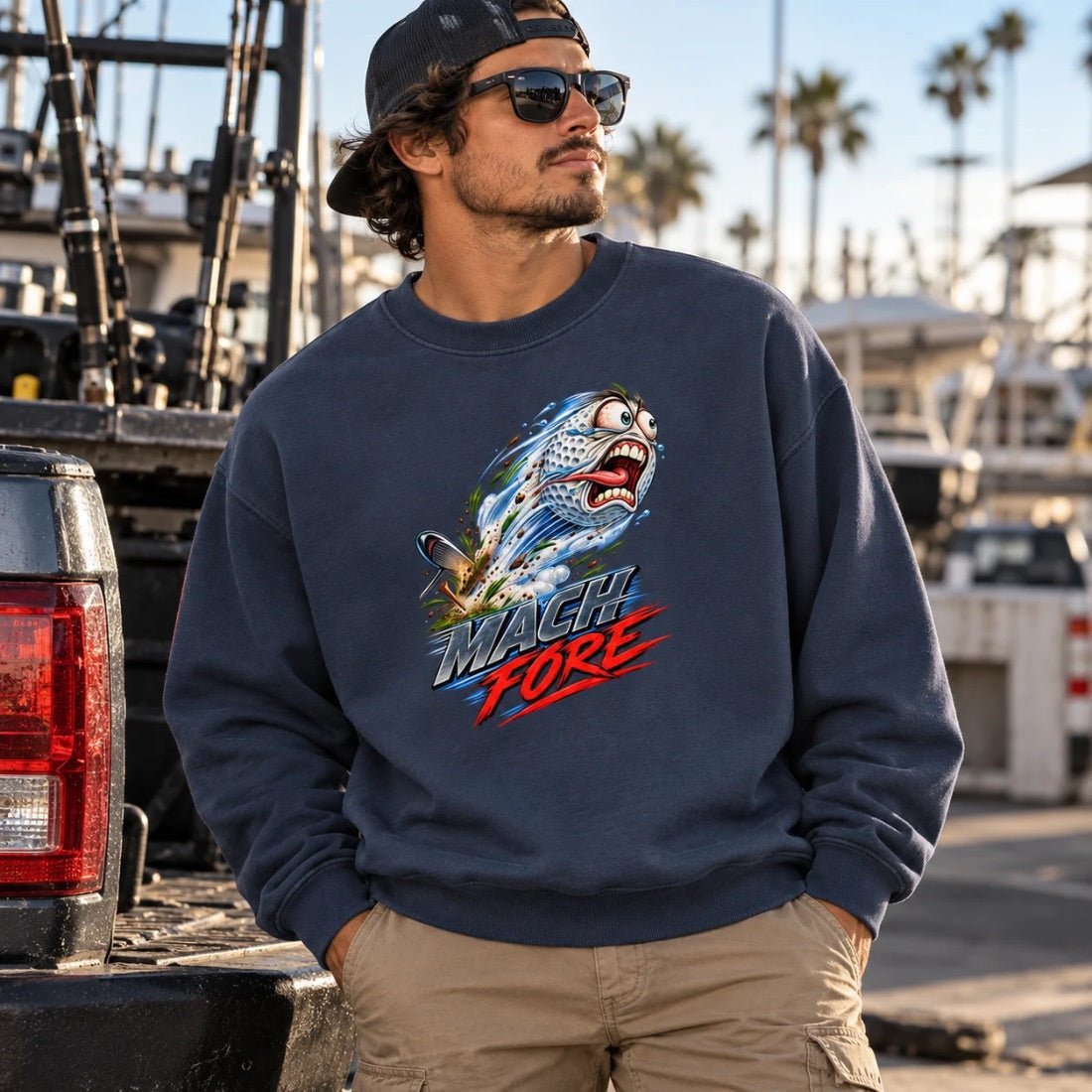 Model wearing a navy blue Mach Fore sweatshirt with a flying golf ball graphic beside a pickup truck.